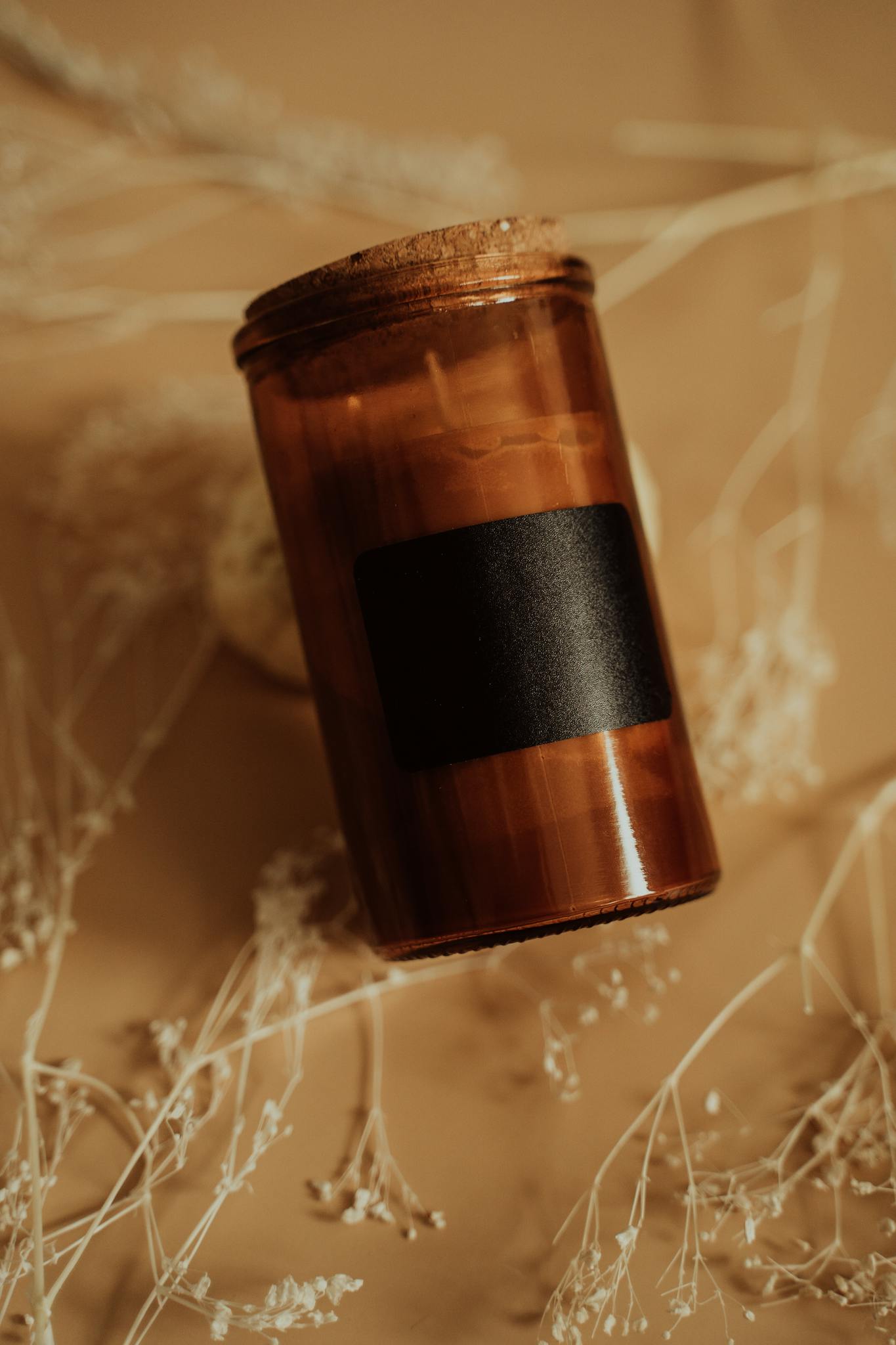 An elegant brown candle with a black label, surrounded by dried flowers on a tan background.